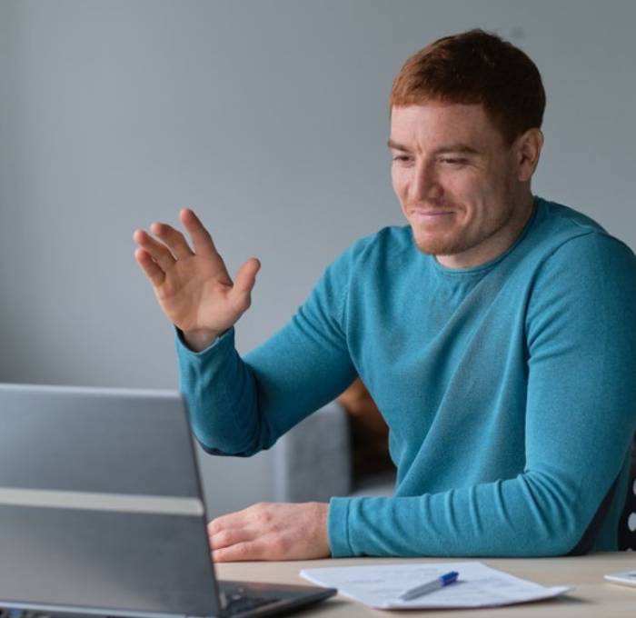 Man zwaait naar het scherm van een laptop tijdens een videogesprek, met papieren en pen op tafel
