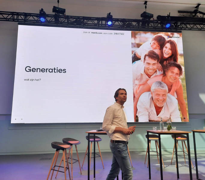 Spreker in een zaal voor een scherm met de slide Generaties, wat zijn het? en een familiefoto