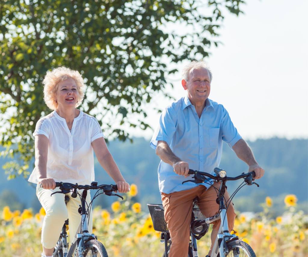 Twee oudere personen fietsen lachend langs een veld met zonnebloemen op een zonnige dag