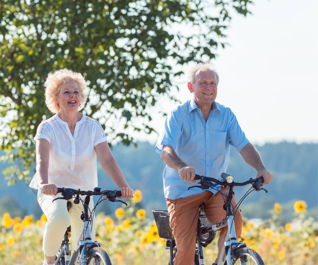 Twee oudere personen fietsen lachend langs een veld met zonnebloemen op een zonnige dag