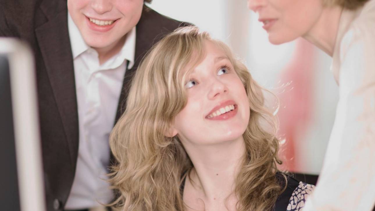 Jonge vrouw kijkt glimlachend op naar twee mensen die met haar meekijken bij een beeldscherm