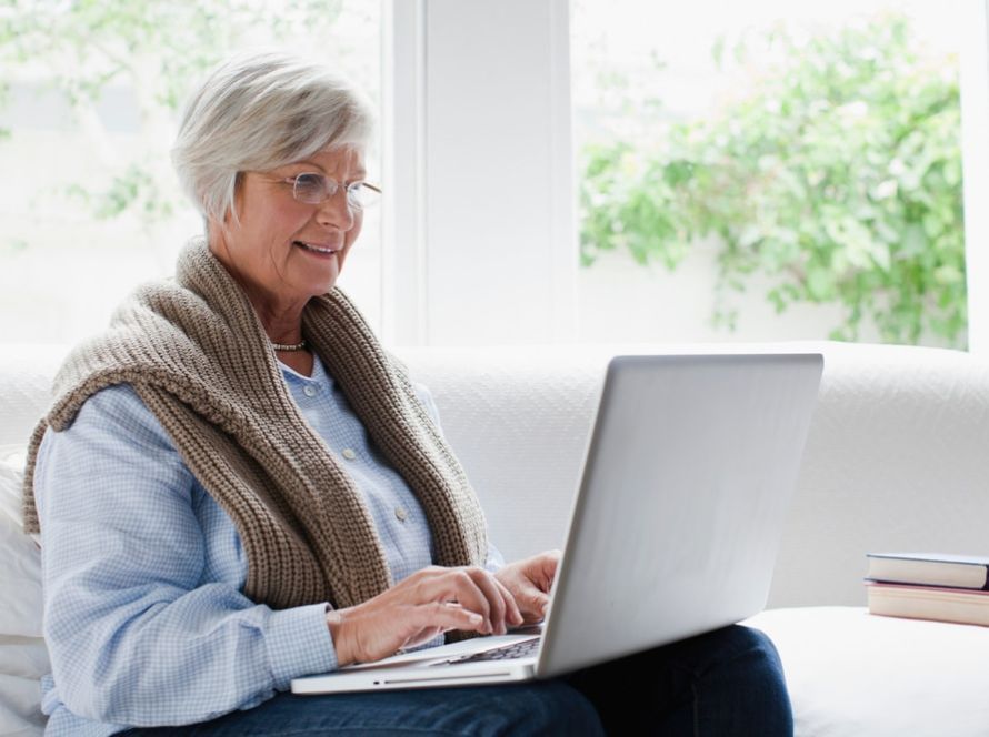 Oudere vrouw met bril werkt op een laptop op de bank bij een raam