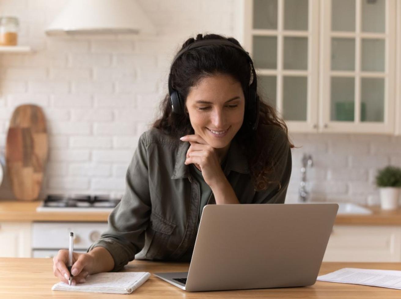 Vrouw met koptelefoon maakt aantekeningen tijdens een online sessie achter haar laptop aan de keukentafel
