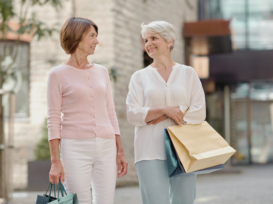 Twee vrouwen lopen met winkeltassen door een winkelstraat en kijken elkaar lachend aan
