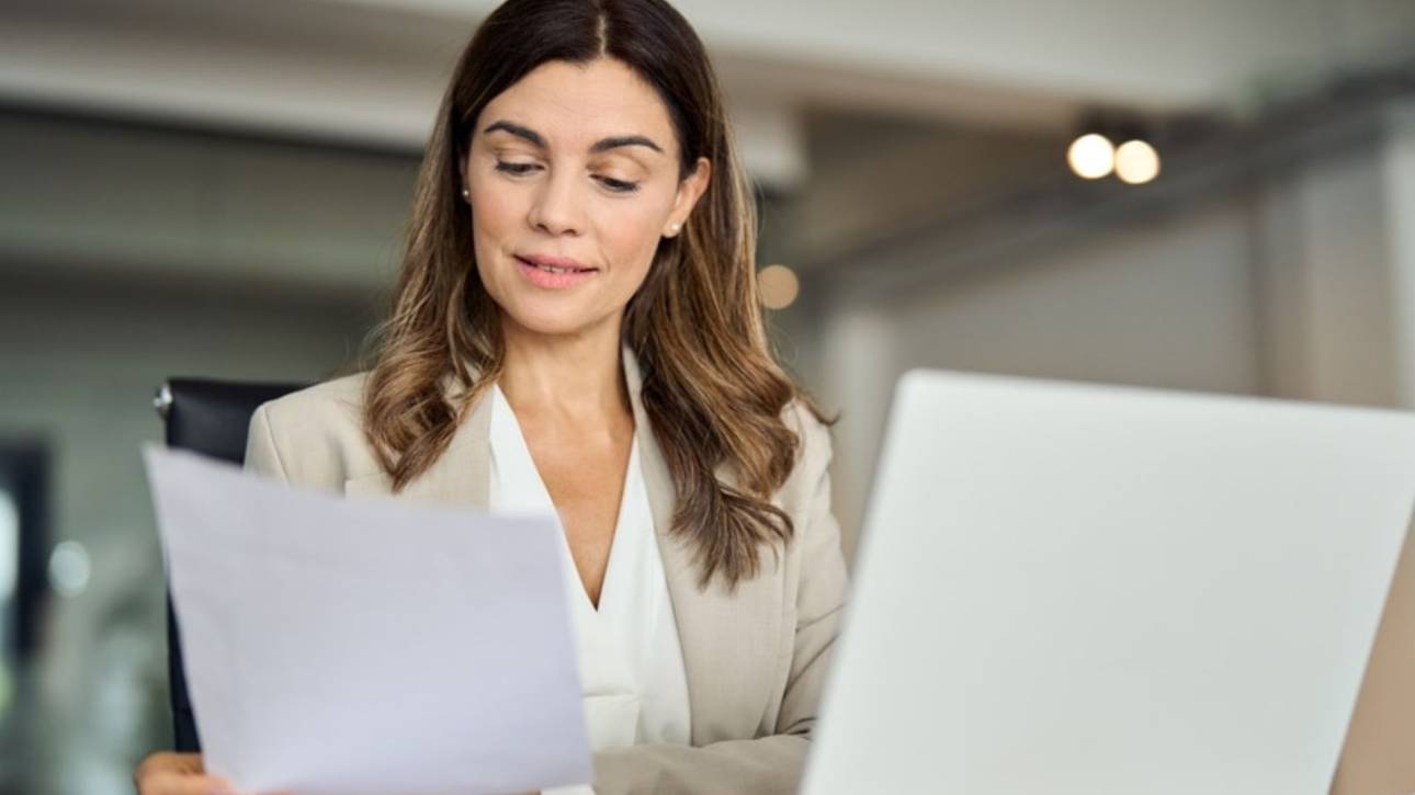 Persoon in blazer leest een document naast een laptop op kantoor