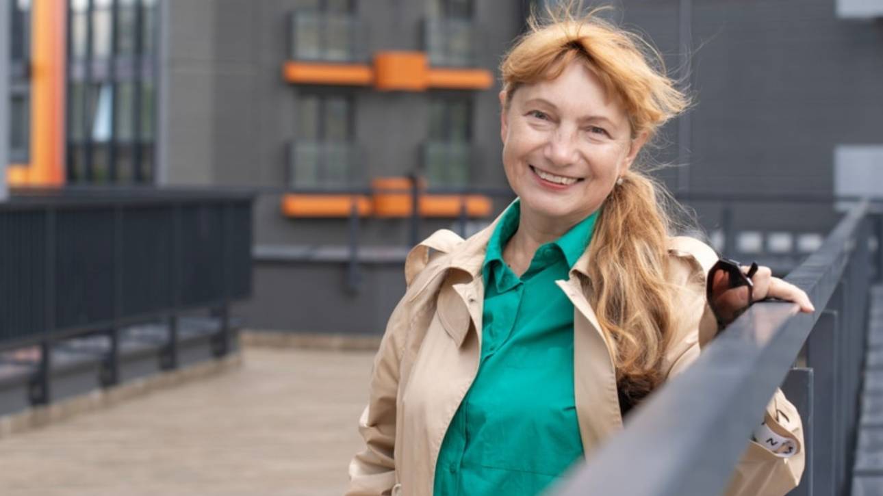 Lachende vrouw in trenchcoat en groene blouse staat buiten bij een leuning voor een modern gebouw