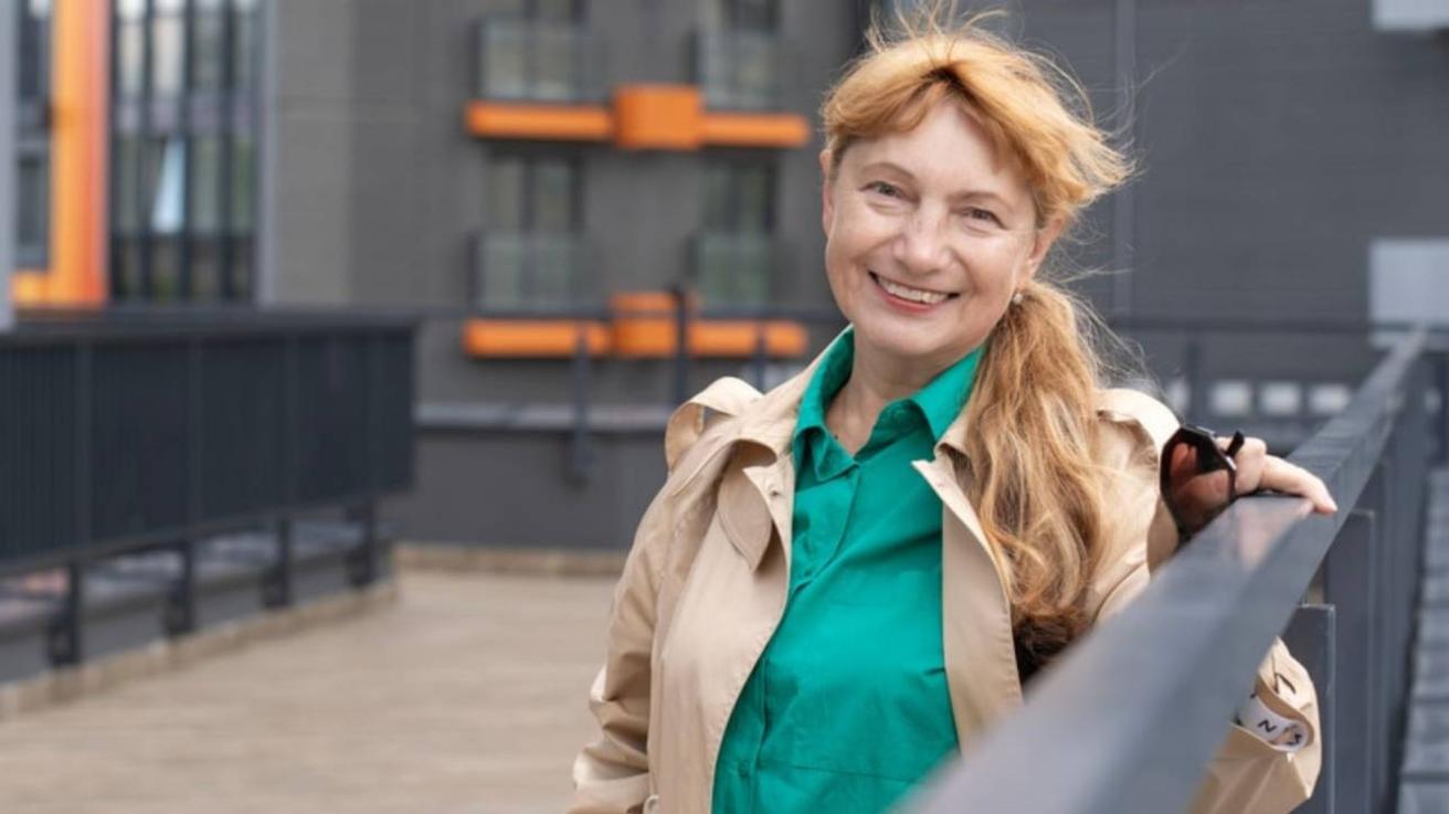 Lachende vrouw in trenchcoat en groene blouse staat buiten bij een leuning voor een modern gebouw