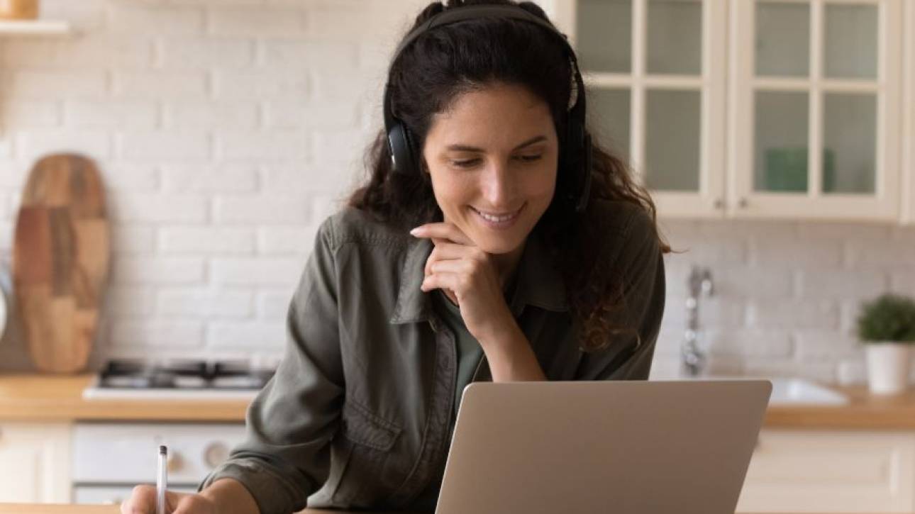 Persoon met koptelefoon volgt lachend een online sessie achter een laptop en maakt aantekeningen