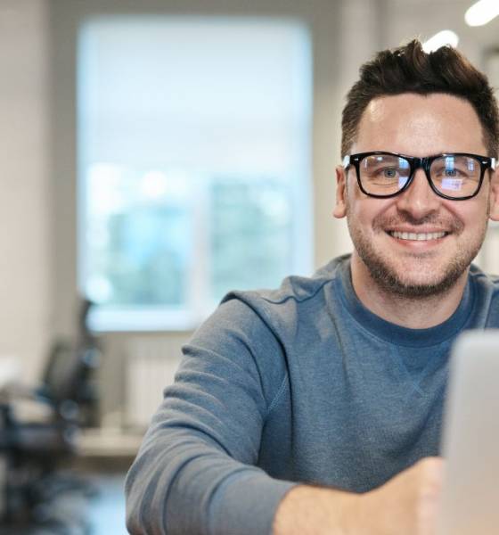 Lachende persoon met bril zit achter een computerscherm in een open kantoorruimte