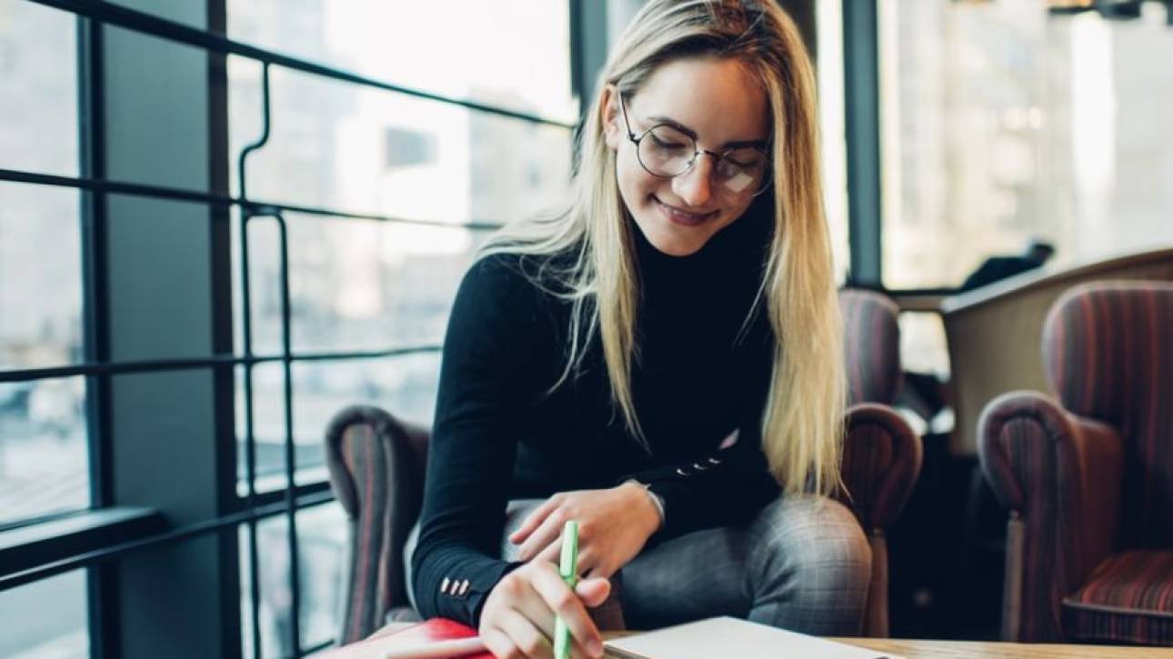 Vrouw met bril schrijft in een notitieboek aan een tafel bij het raam van een café