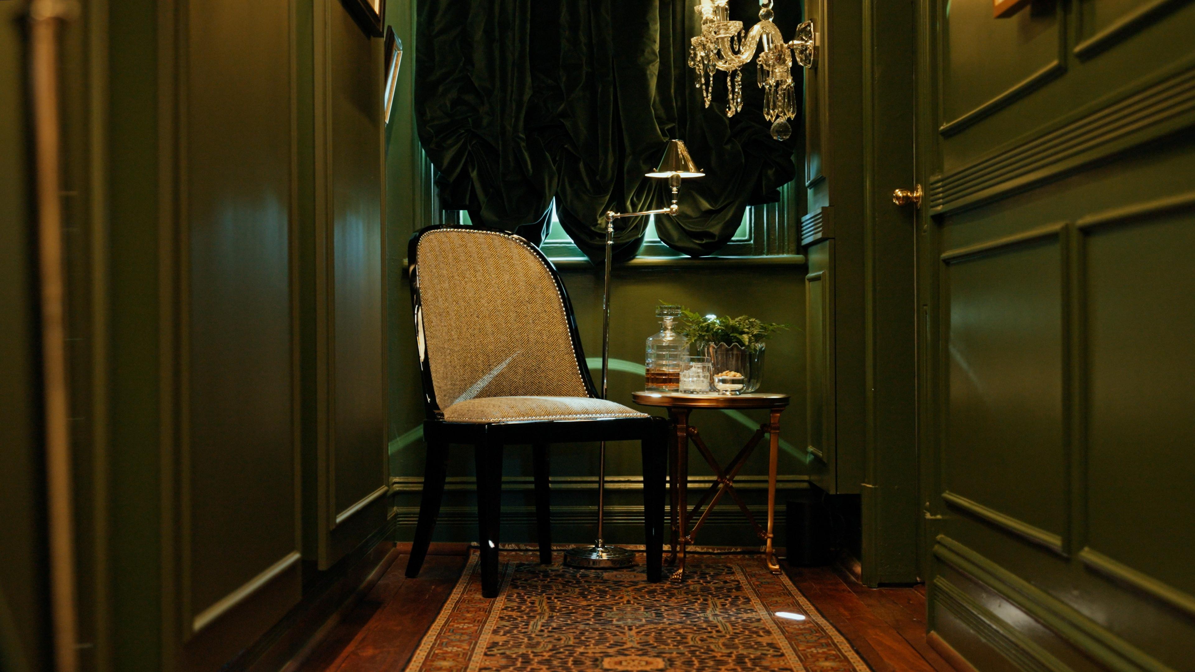 An upholstered chair at the end of a green panelled hallway, beneath a crystal chandelier