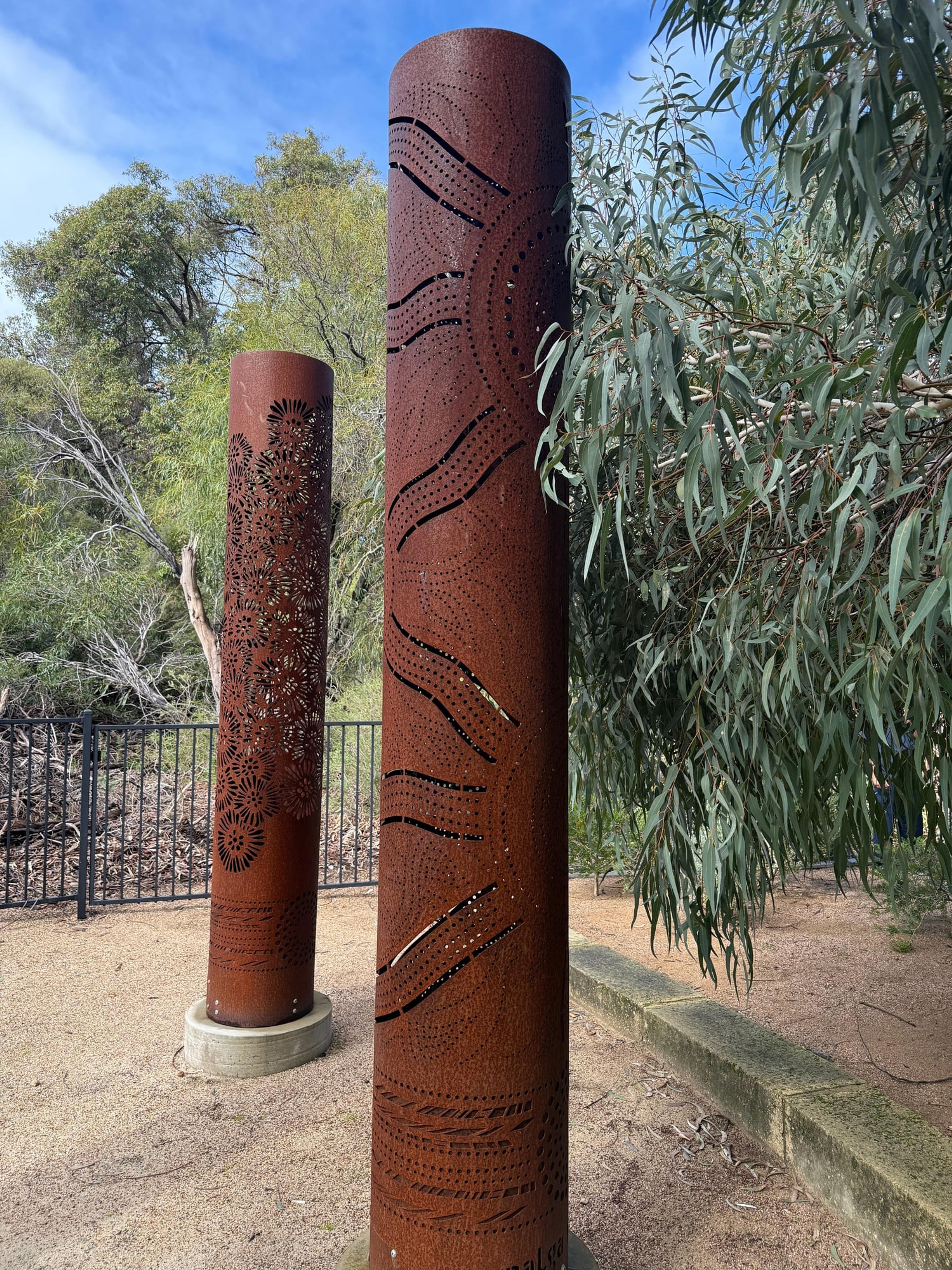 Two rusted totems, one with wavy cut lines and one with a floral pattern, beside gum leaves