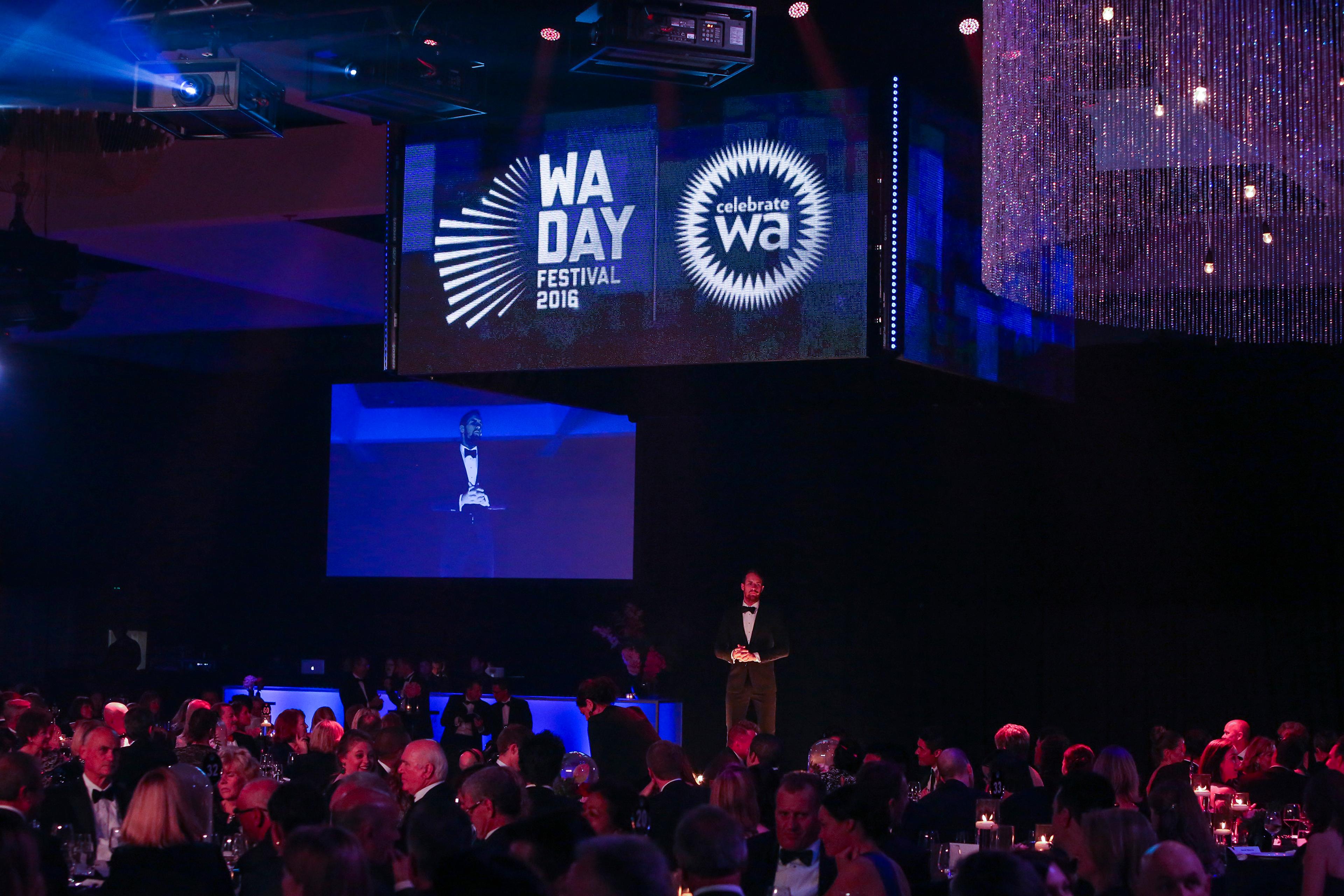 The host on stage at the Western Australian of the Year Awards, under the WA Day Festival screens