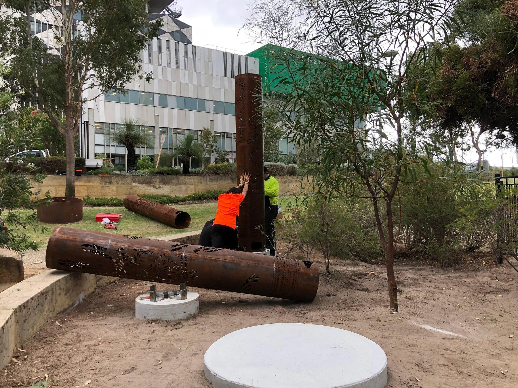 Installers lifting a totem into place with the hospital behind