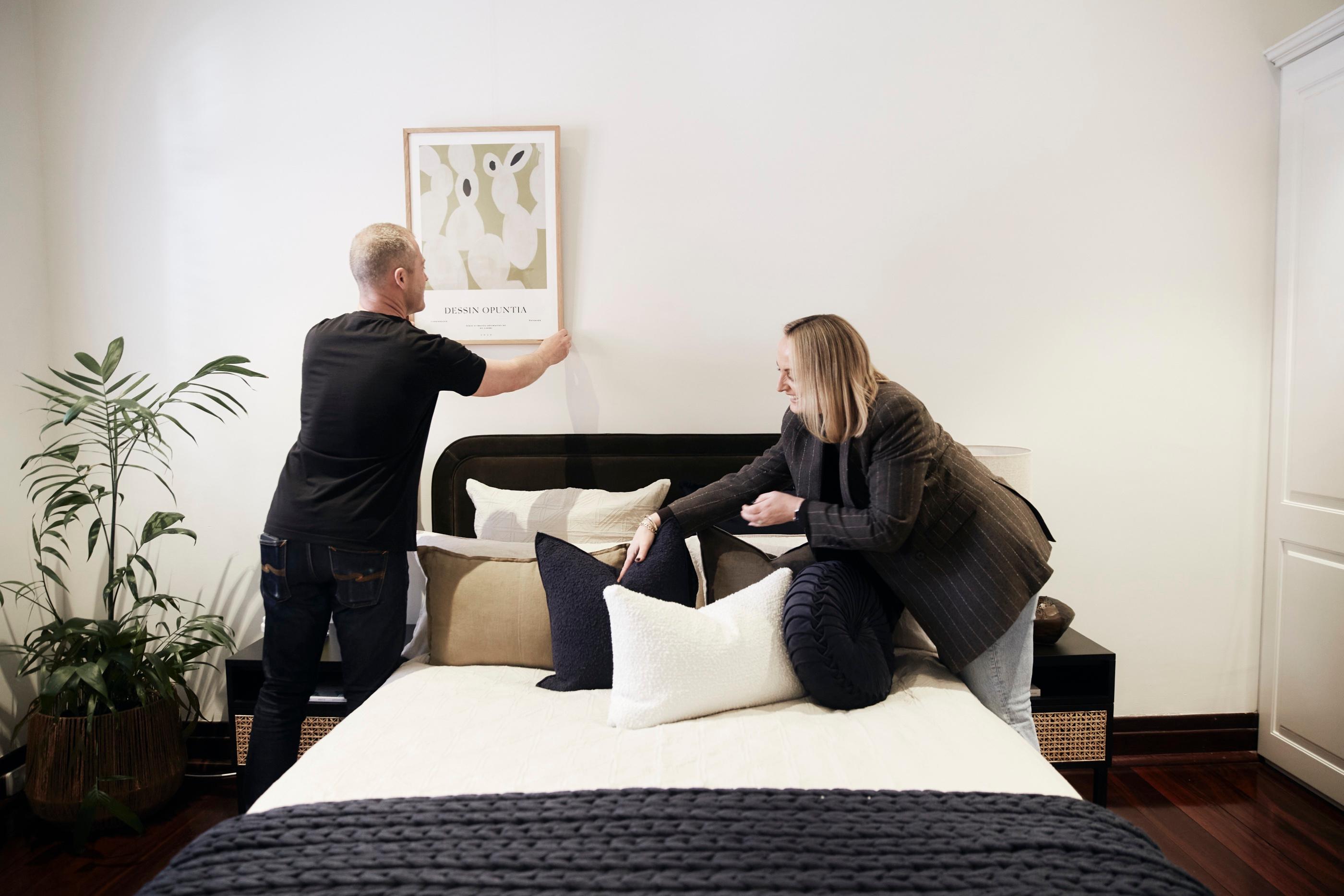 Dan hanging art while Nadija styles the cushions on a bed