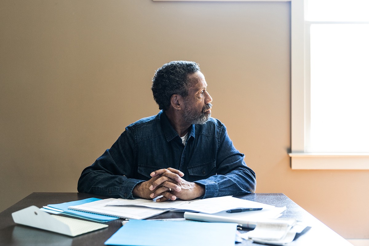 An older man with gray hair sits at a kitchen table covered in papers, folders and notebooks. He looks to his left out of of frame.