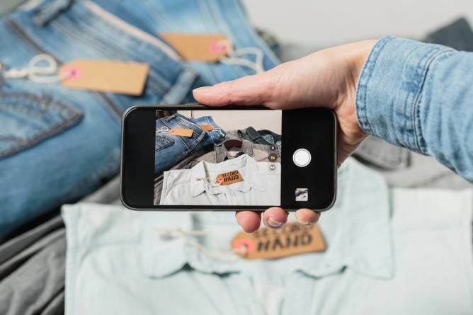 A woman's hand holds a smartphone while taking a photograph of a shirt with a tag that says "secondhand."