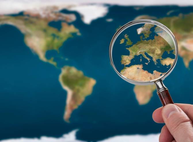 simple map of the world; in the foreground a hand holds a magnifier over Europe and Northern Africa