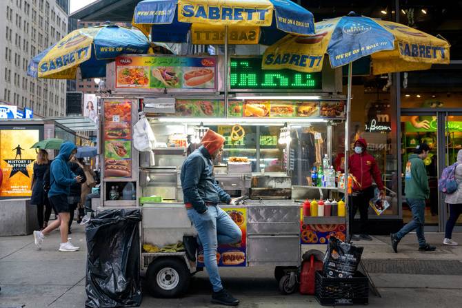 A halal cart worker checks his phone as people walk past across the road from the unveiling on The Broadway Grand Gallery in Times Square