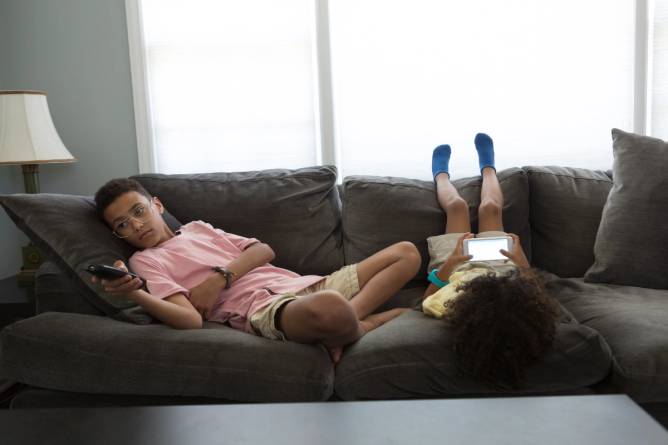 Two boys sit on a gray couch, one holding a remote and the other looking at the screen of a mobile phone 