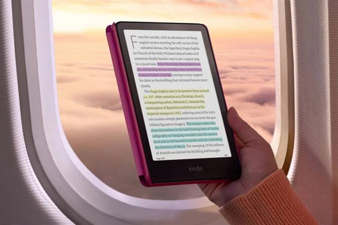 A hand holding the Kindle Colorsoft in front of an airplane window