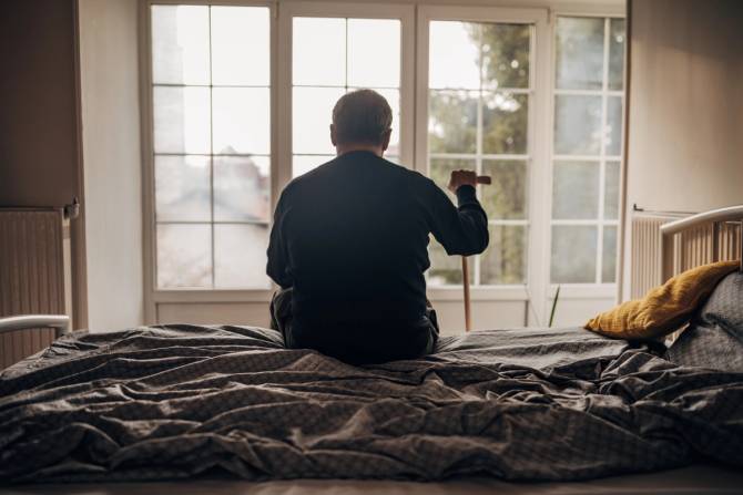 A man sits on his bed in a nursing home facing a window.