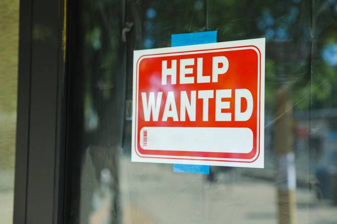 A "help wanted" sign, taped to the glass door of a business establishment.