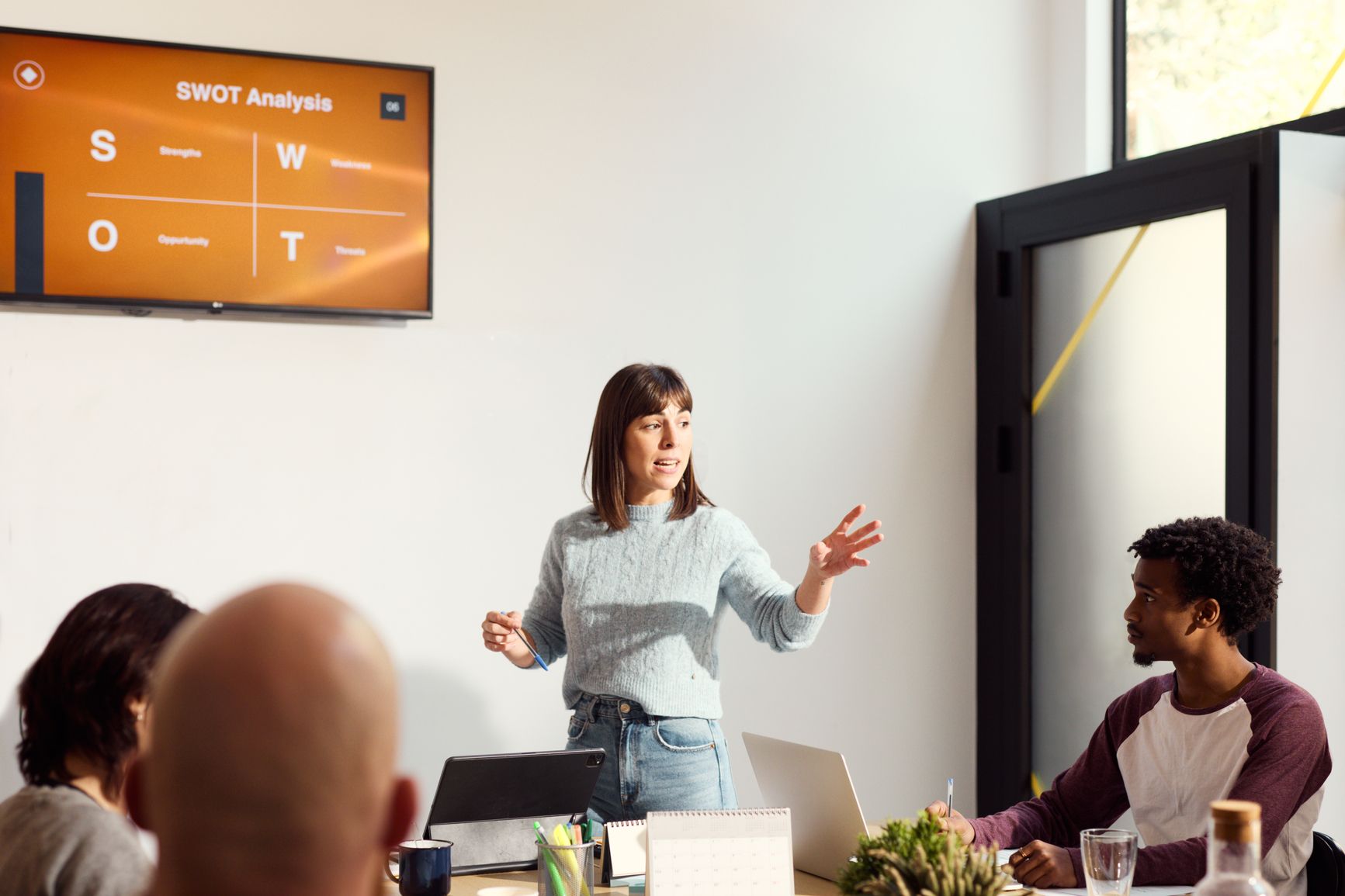 A woman presents a SWOT analysis slide on a wall-mounted screen to three colleagues seated around a conference table.