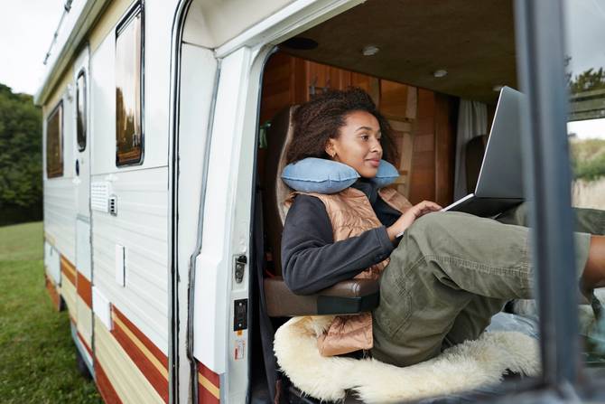 A person with a neck pillow sits in a camper van with a laptop computer.