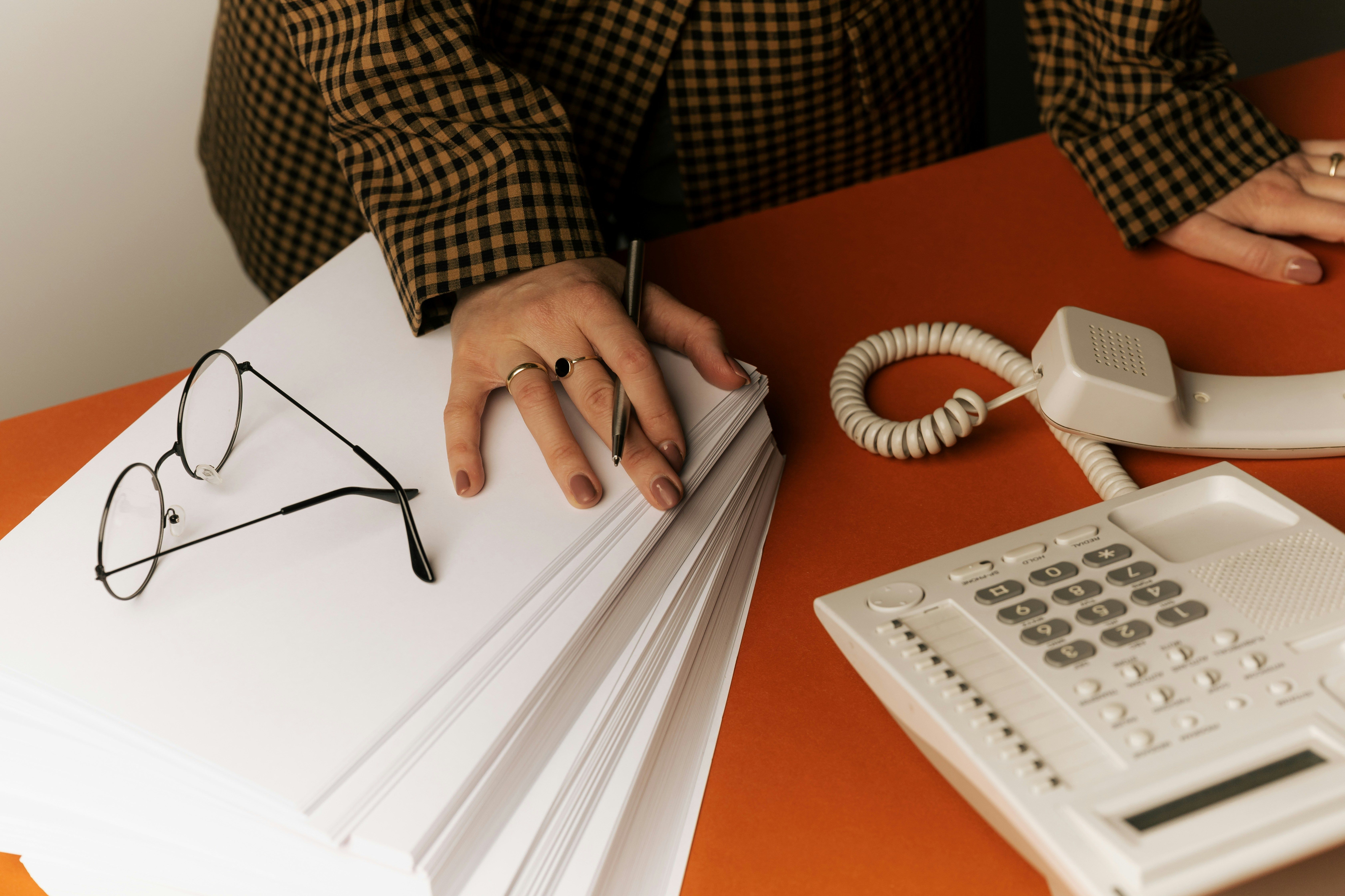 A person's hands resting on a stack of papers on an orange desk, with round wire-framed glasses and a white desk telephone nearby.