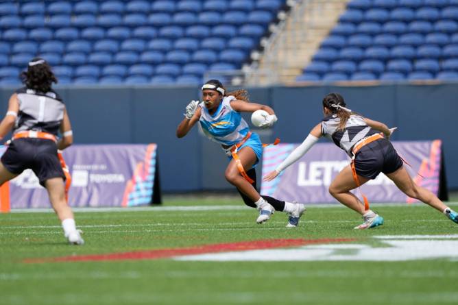 A woman holding a football in her arms runs towards two other women facing her on a field.
