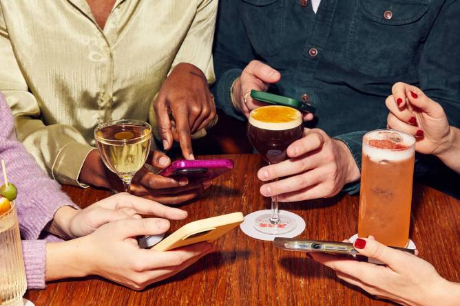 a photograph of four pairs of hands holding phones over a dining table with four cocktail glasses containing drinks