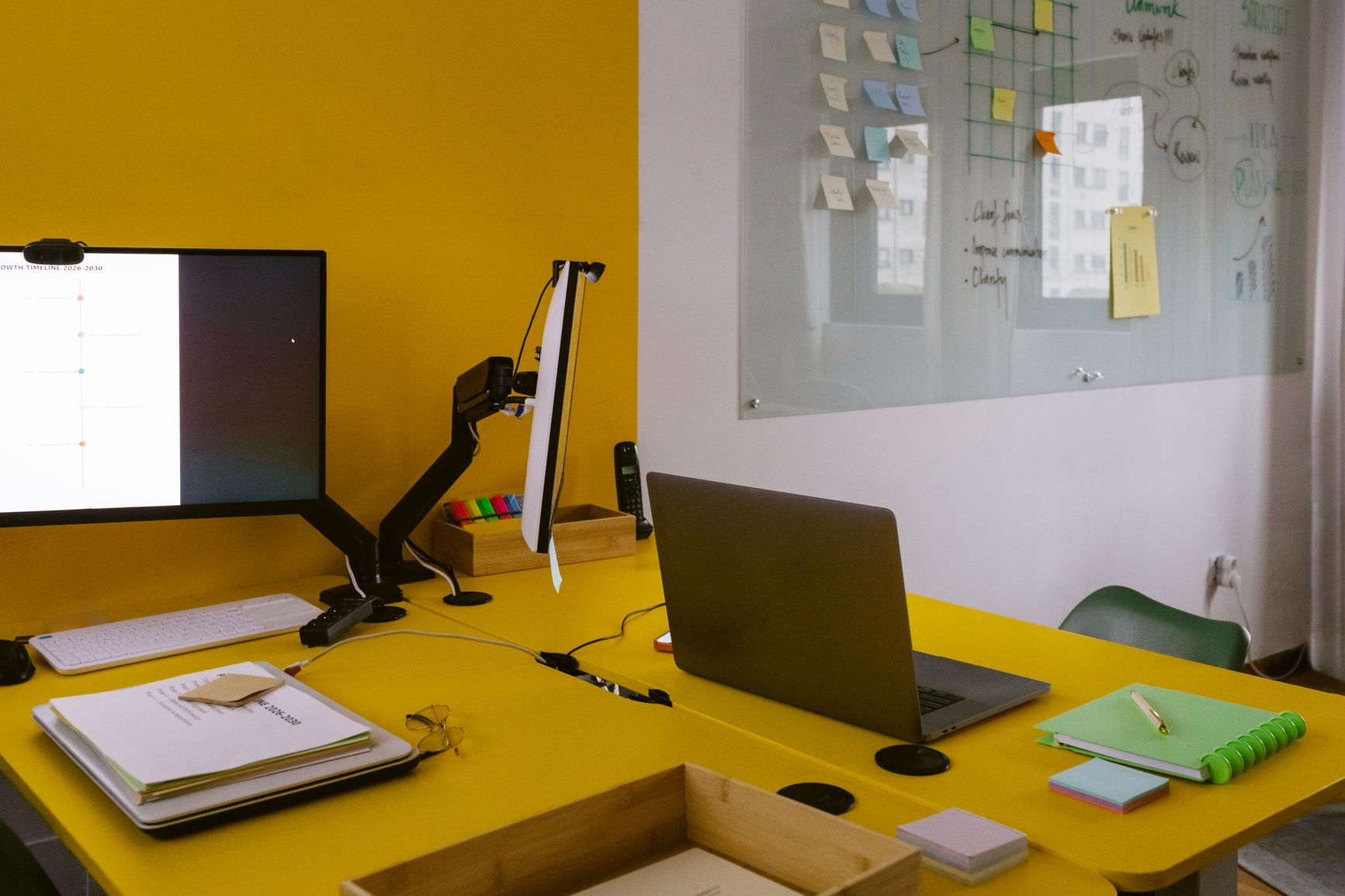 A bright yellow office workspace featuring a dual-monitor setup on a monitor arm, an open laptop, scattered notebooks and sticky notes, and a glass whiteboard covered in colorful sticky notes and handwritten diagrams on the wall behind.