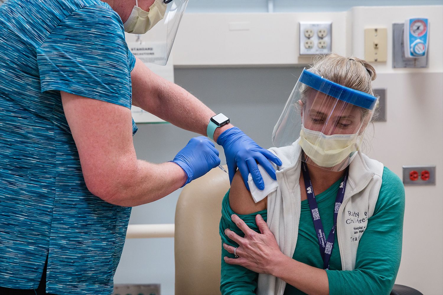 A nurse administers a Covid-19 vaccine shot to a doctor, both are wearing face masks and face shields.