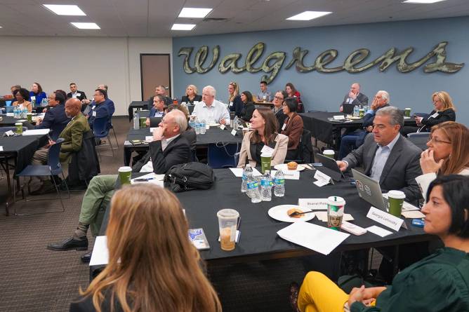 A room full of people sitting at tables with a Walgreens logo behind them