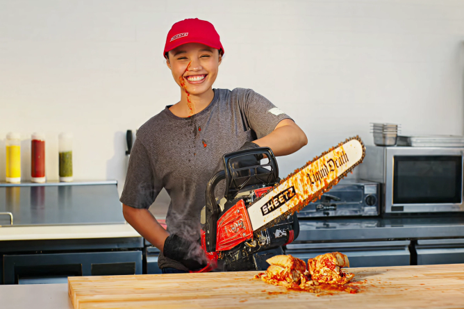 A woman holds a Liquid Death X Sheets branded chainsaw that she's just cut a sandwich with and it's splattered sauce on her face.