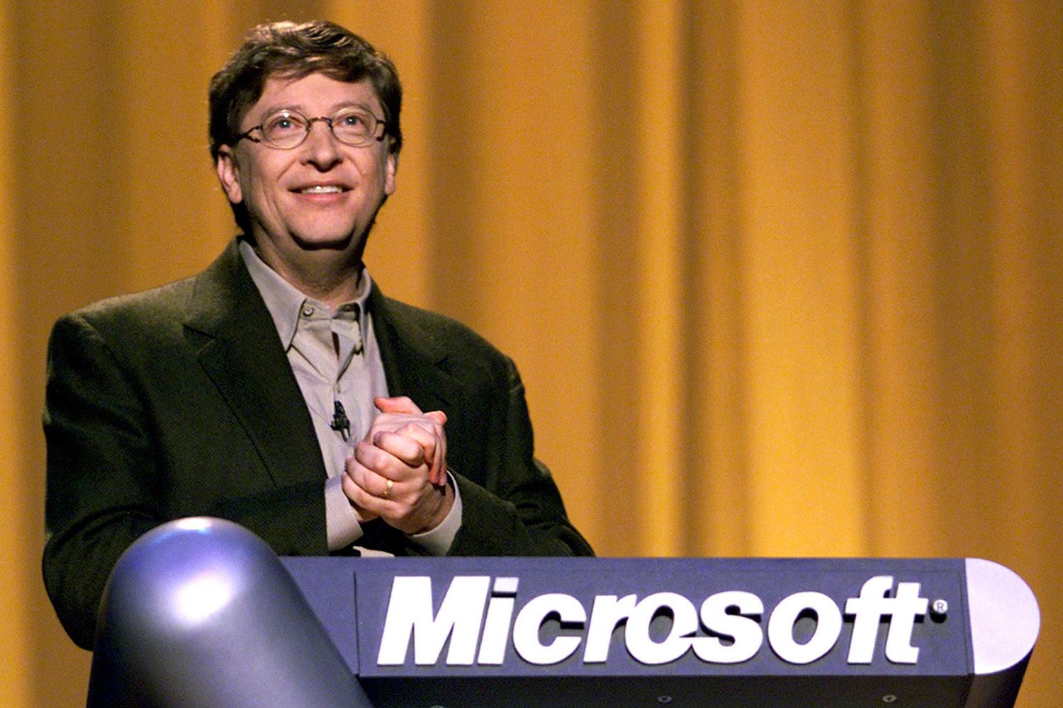 A young Bill Gates in a suit stands clasping his hands in front of a podium with the Microsoft logo on the front.