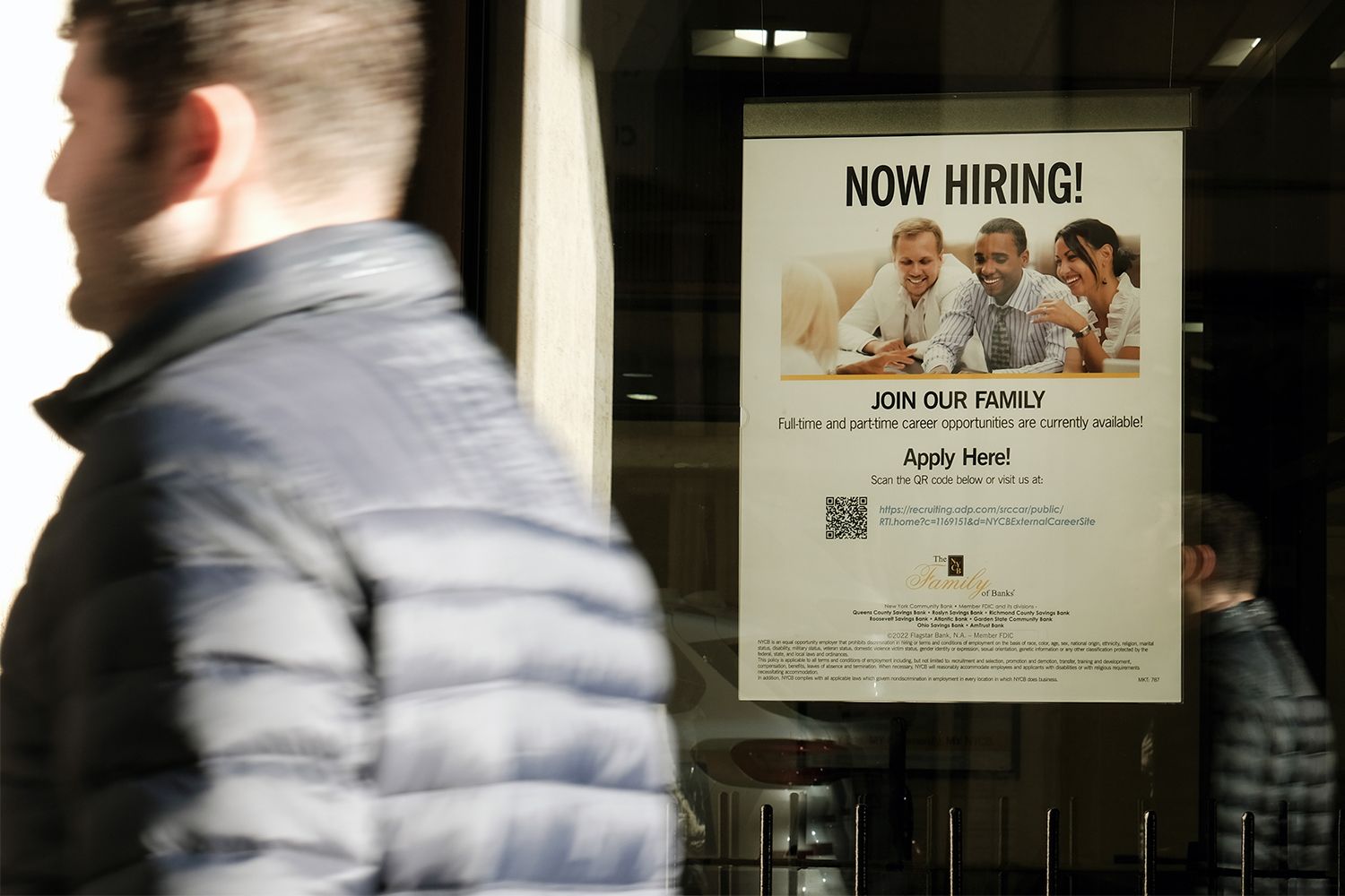 A man in the foreground walks past a Now Hiring sign on a storefront window.