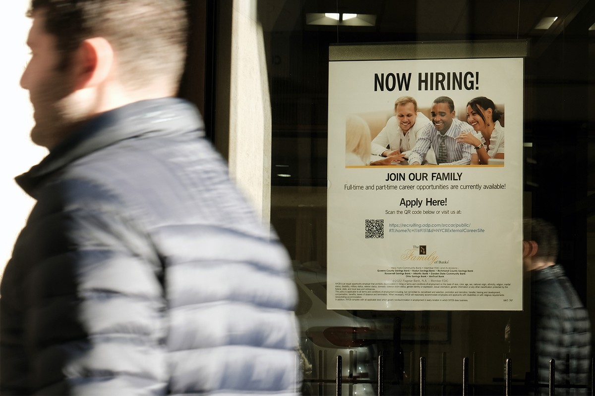 A man in the foreground walks past a Now Hiring sign on a storefront window.