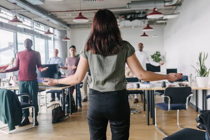 A woman standing in front of her colleagues with he arms outstretched, leading them in group meditation at the office.