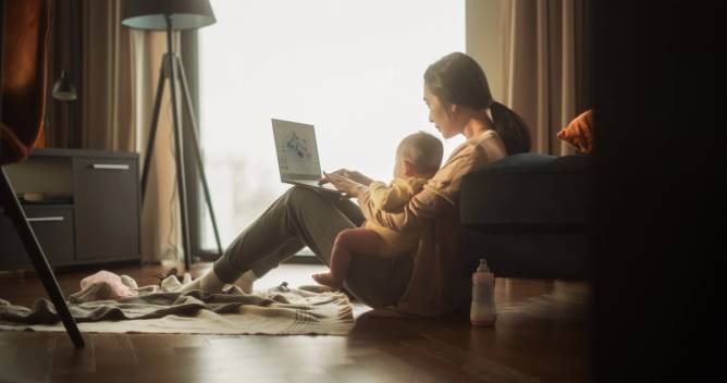 A woman sits on the floor and works from her laptop while holding a baby in her lap.