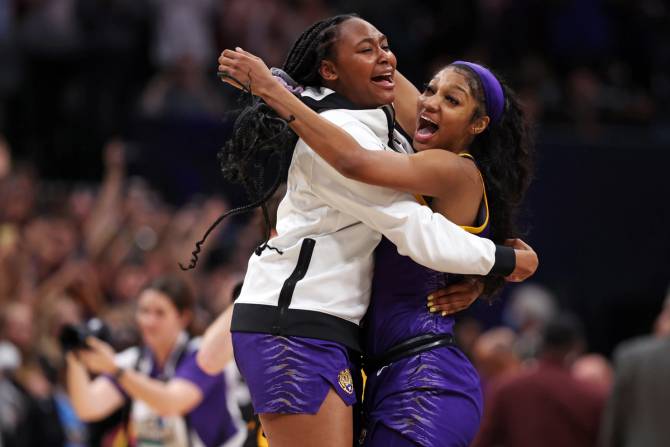 A close-up photo of Angel Reese of the LSU Lady Tigers, wearing purple shorts and a purple headband, embracing a teammate wearing purple shorts and a white pullover, with spectators and a photographer in the background.