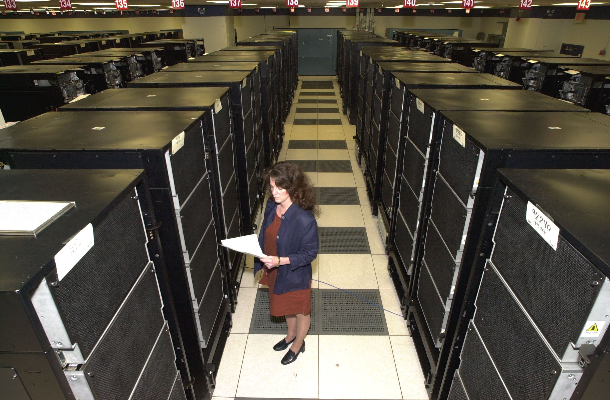 A woman in a blazer stands in a hallway created by computer towers. At least 6 rows of similar towers are visible in the room.