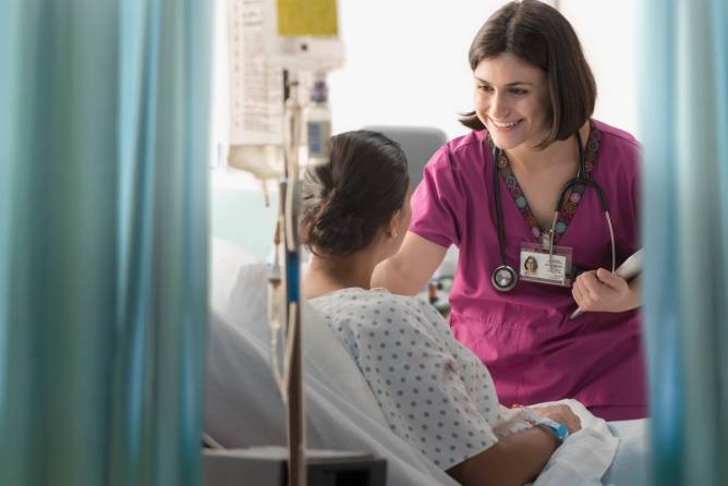 A female nurse in magenta scrubs speaks to a patient.
