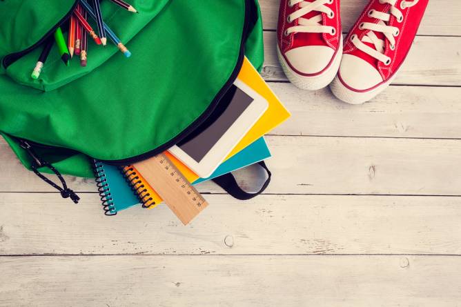 Image of a green backpack with school supplies and a tablet against a wooden background.