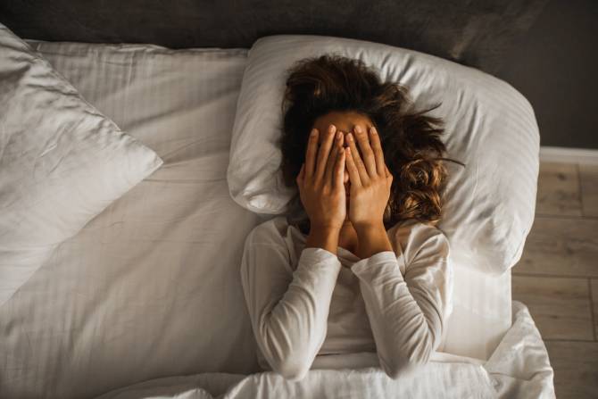 A woman lays in bed with her hands over her face in despair.