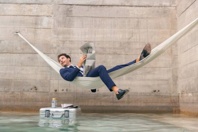 Man reading newspaper in a hammock suspended over water with with a beer on top of a floating briefcase.