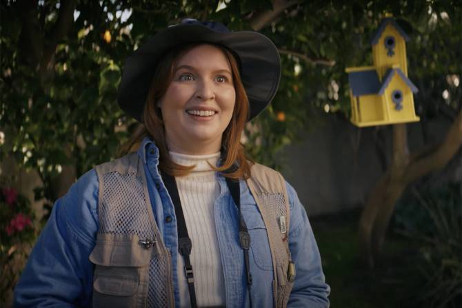 a woman in a jean jacket and hat staring at a yellow birdhouse outside