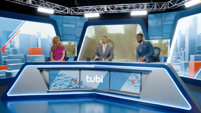 Three sports anchors—a woman in a pink dress and two men in suits—sit on a sports commentary-like set engaged in dialogue behind a desk with the Tubi logo on it.