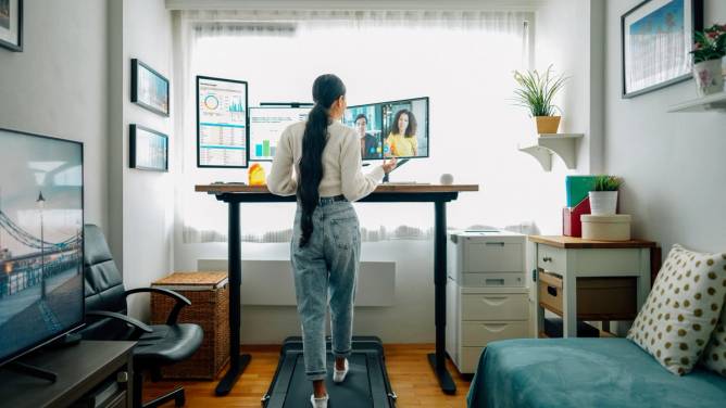 Woman walking on a treadmill in front of a desk while taking a video call.